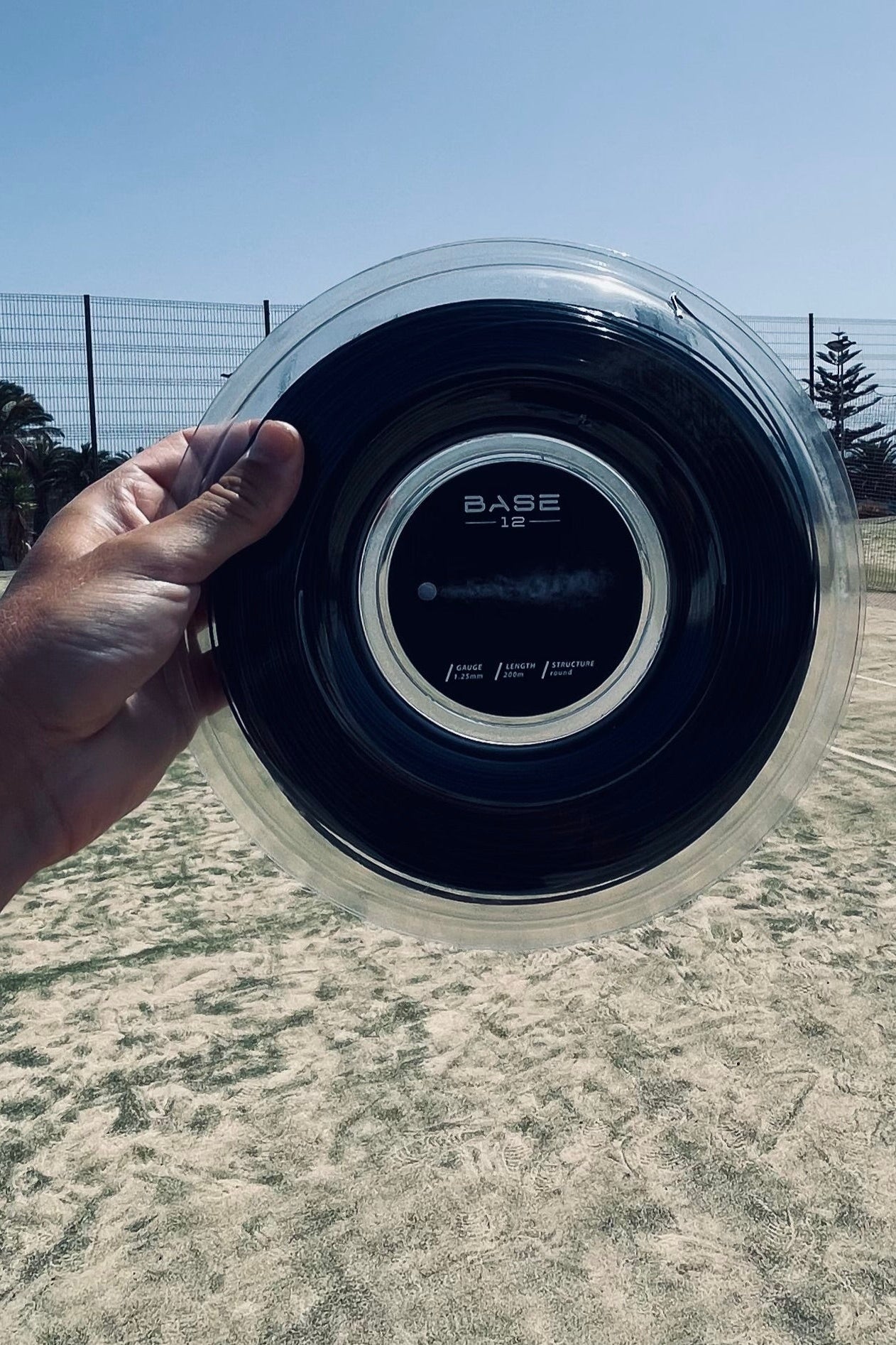 Tennissaite von BASE12 in der Verpackung, gehalten auf einem Tennisplatz unter freiem Himmel.