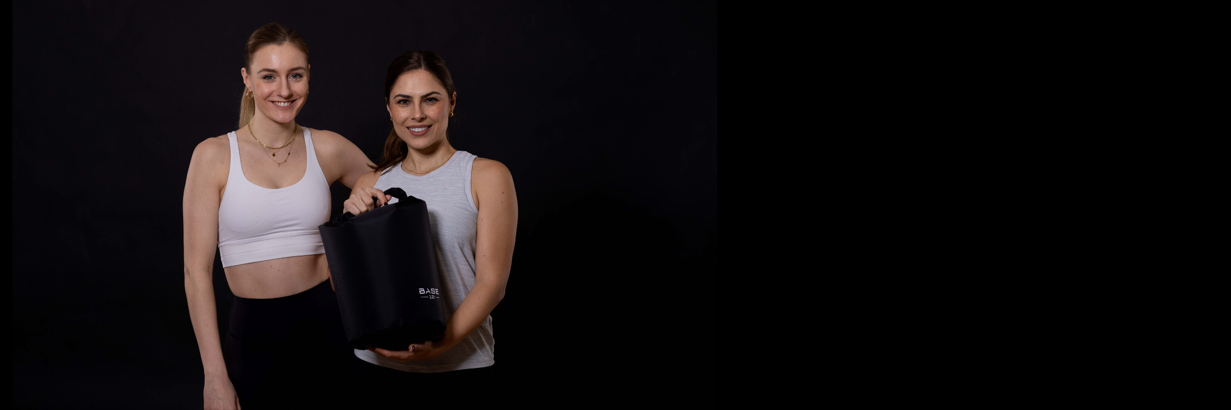 Zwei Tennisspielerinnen stehen nebeneinander und halten eine BASE12 Sporttasche – lächelnd und bereit für das Training