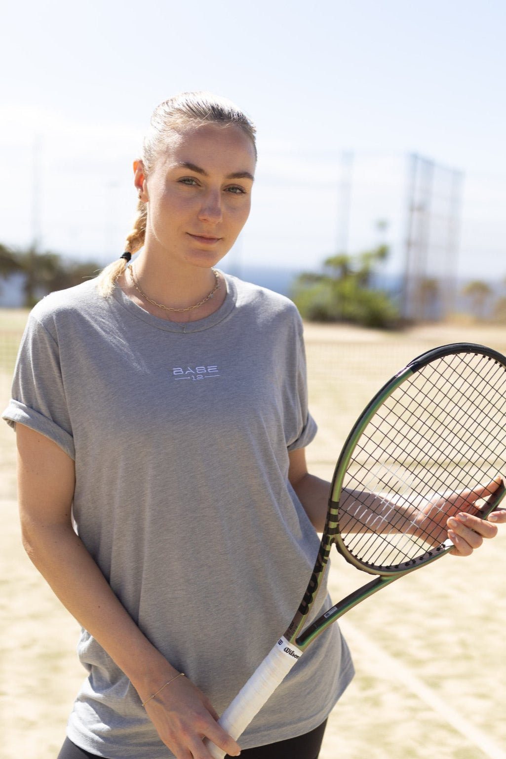 Tennisspielerin trägt ein graues BASE12 Shirt und hält einen Tennisschläger auf dem Platz
